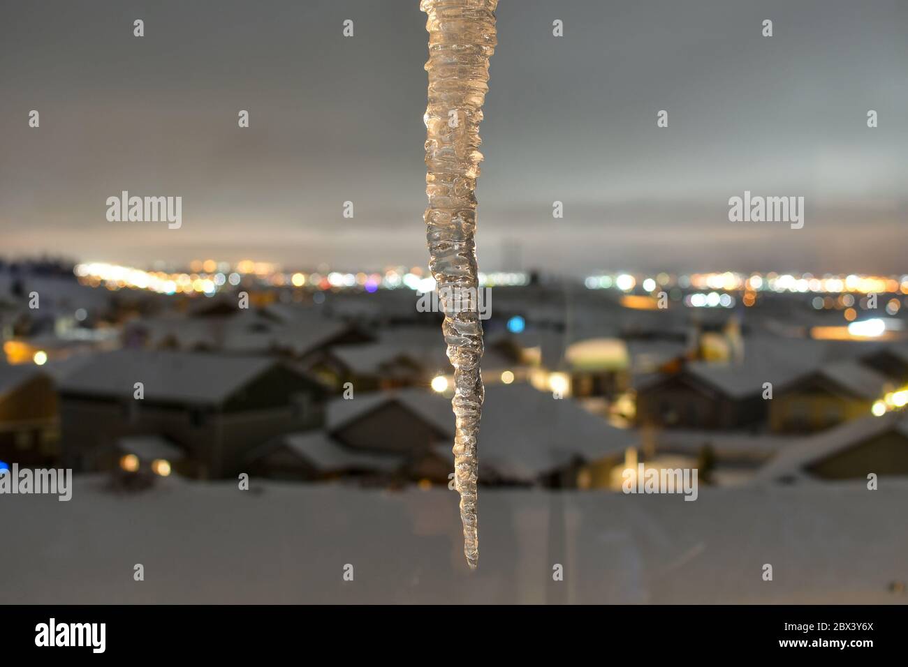 An icicle hangs in front of a blurred view of a subdivision neighborhood, city and mountains in Spokane, Washington covered with snow in winter Stock Photo