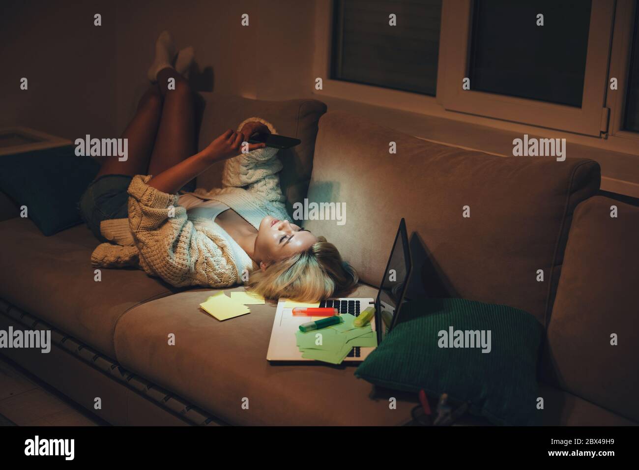Tired caucasian student chatting on phone while sitting on sofa after doing homework late evening Stock Photo