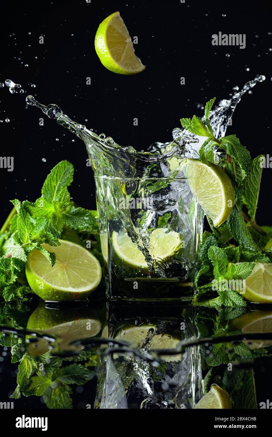 Cocktail with limes and mint on a black reflective background. Lime slice fall in glass with drink. Stock Photo