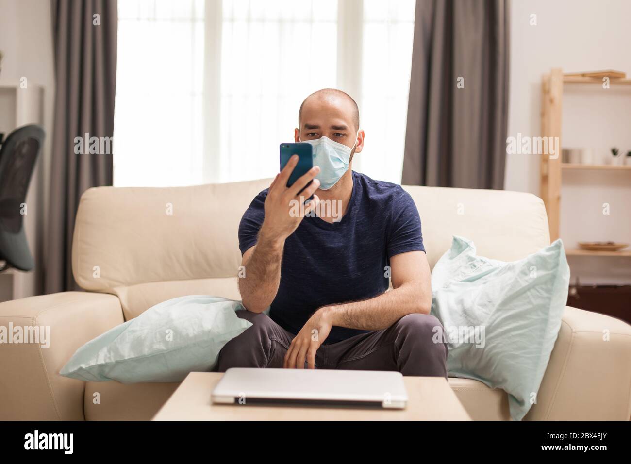 Man watching a video on smartphone wearing protection man while sitting on sofa during quarantine. Stock Photo