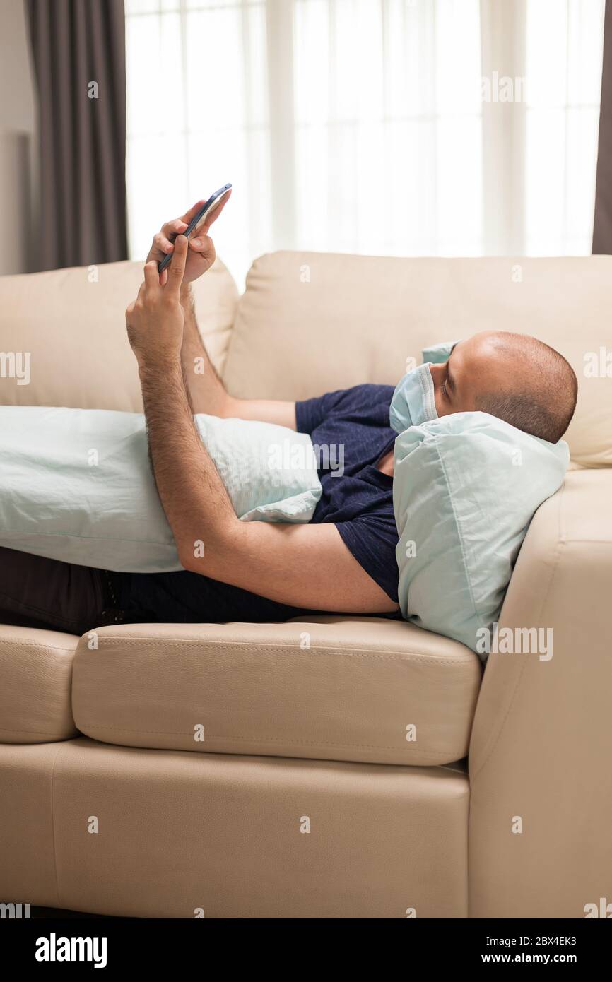 Man laying on sofa using his smartphone during global pandemic. Stock Photo