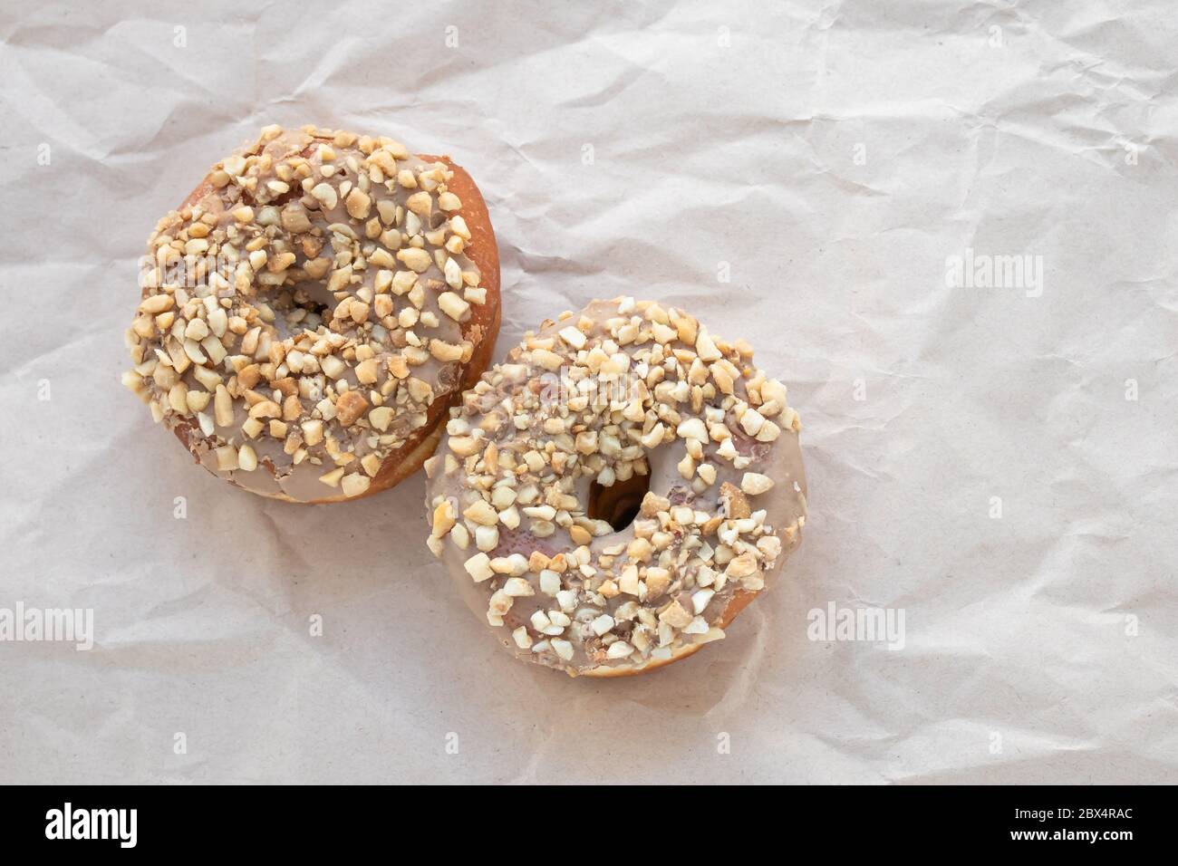 Brown glazed donuts sprinkled with nuts on crumpled beige paper background. Unhealthy high-calorie food. Empty place for text. Copy space Stock Photo