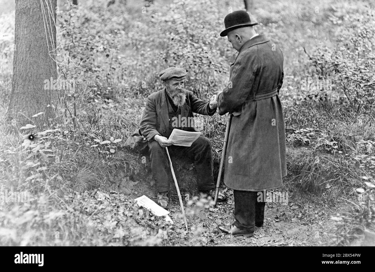 Two men, one sitting, the other standing, look at something they hold in their hands. They both have a stick, one of them is smoking, the other is holding papers in his hand. Further papers are lying on the ground. They are in the Jungfernheide on the way to Saatwinkel, north of the Ploetzensee Canal in the forest.  There, so-called 'monarchs' live in old 'Stubbenloecher' (holes of dear tree trunks), who only possess about 3 sqm large soil. . Due to the world economic crisis and mass unemployment, many people lost their homes. Stock Photo