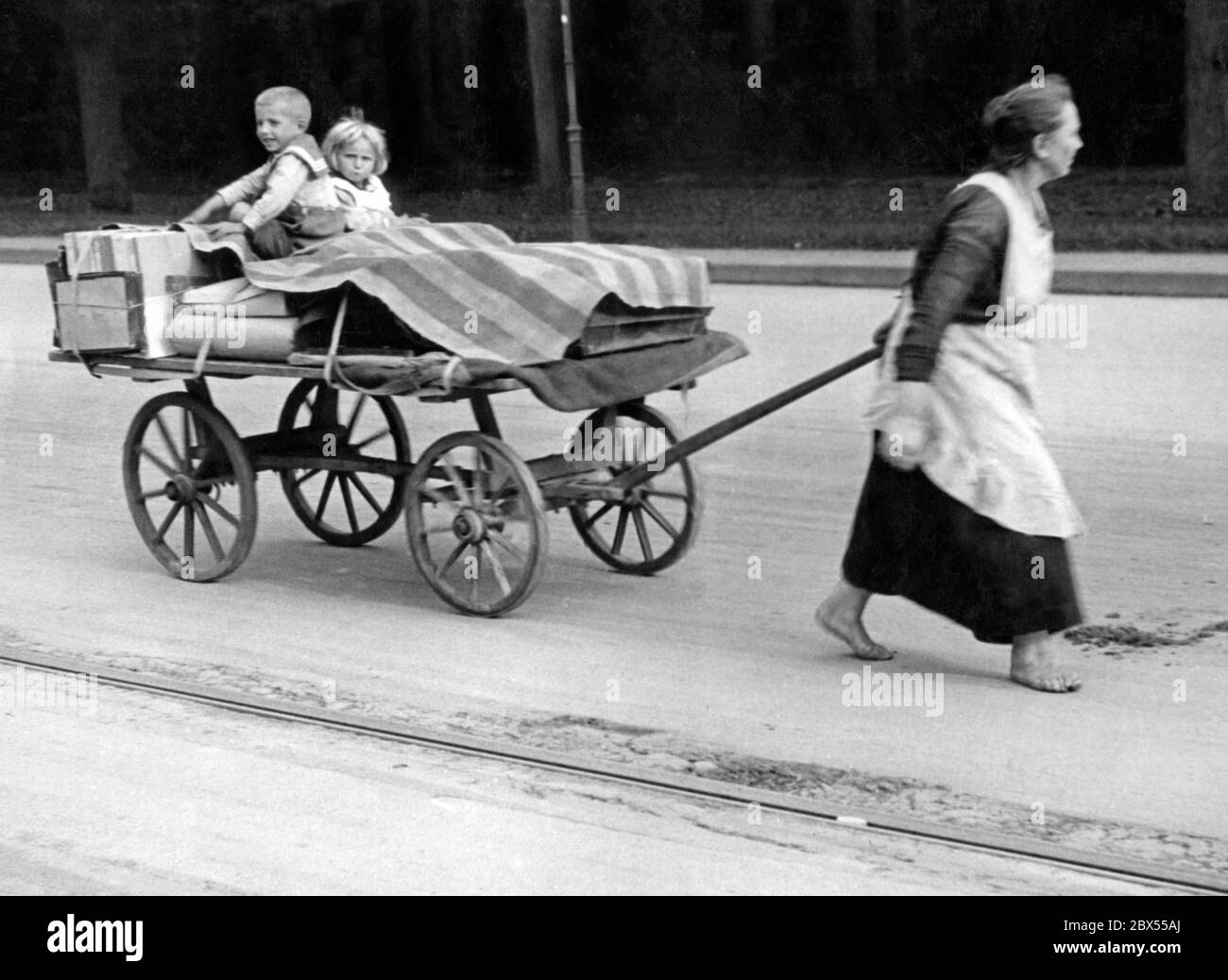 A barefoot woman from a working-class neighbourhood in Berlin, pulls a large cart loaded with some goods under a tarpaulin as well as a boy and a girl. Many working-class women earned a little extra money, sometimes through hard physical labour, as the state's welfare laws were not sufficient. The woman walks barefoot, as she saves her shoes for the winter. Stock Photo