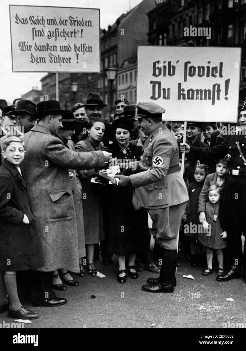 During a street assembly of the Winterhilfswerk des Deutschen Volkes (Winter Relief of the German People), organized by the German Labor Front, peace slogans are held up after the Munich conference (The Reich and Peace are secured! We thank and help the Fuehrer). During the fundraising 'Fuehrerbuechlein' are sold for 'As much as you can afford' (poster). Stock Photo