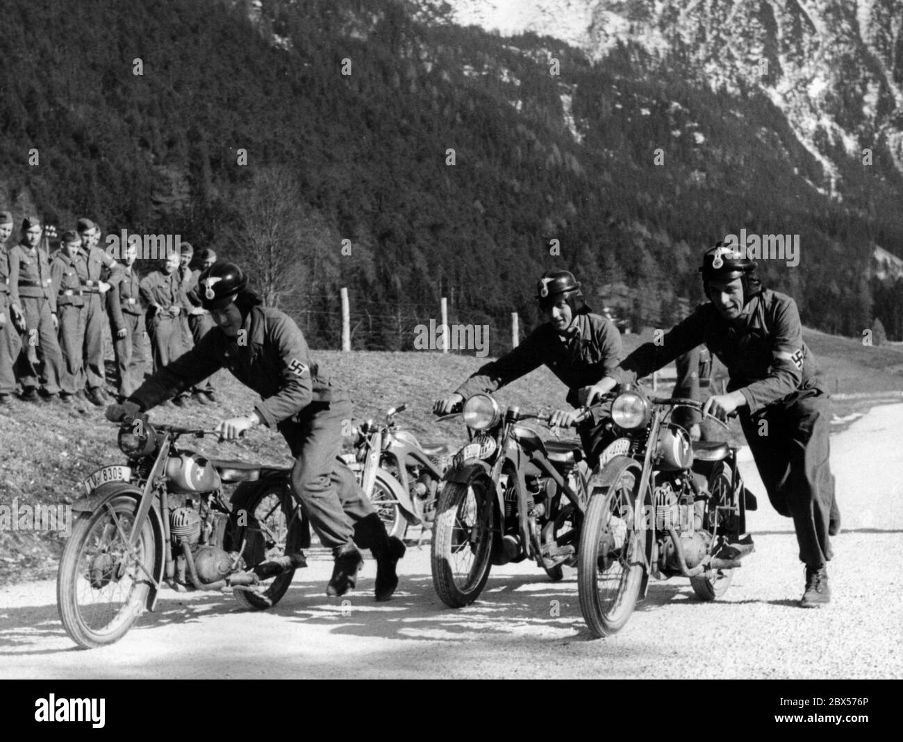 Three members of the Motor Hitler Youth push their motorcycles on a country road for a practice ride. On the left there is a group watching them. In the background are mountains. The motorbike in the middle is a Puch 200. On the left and right are Puch 125 T motorcycles. Stock Photo