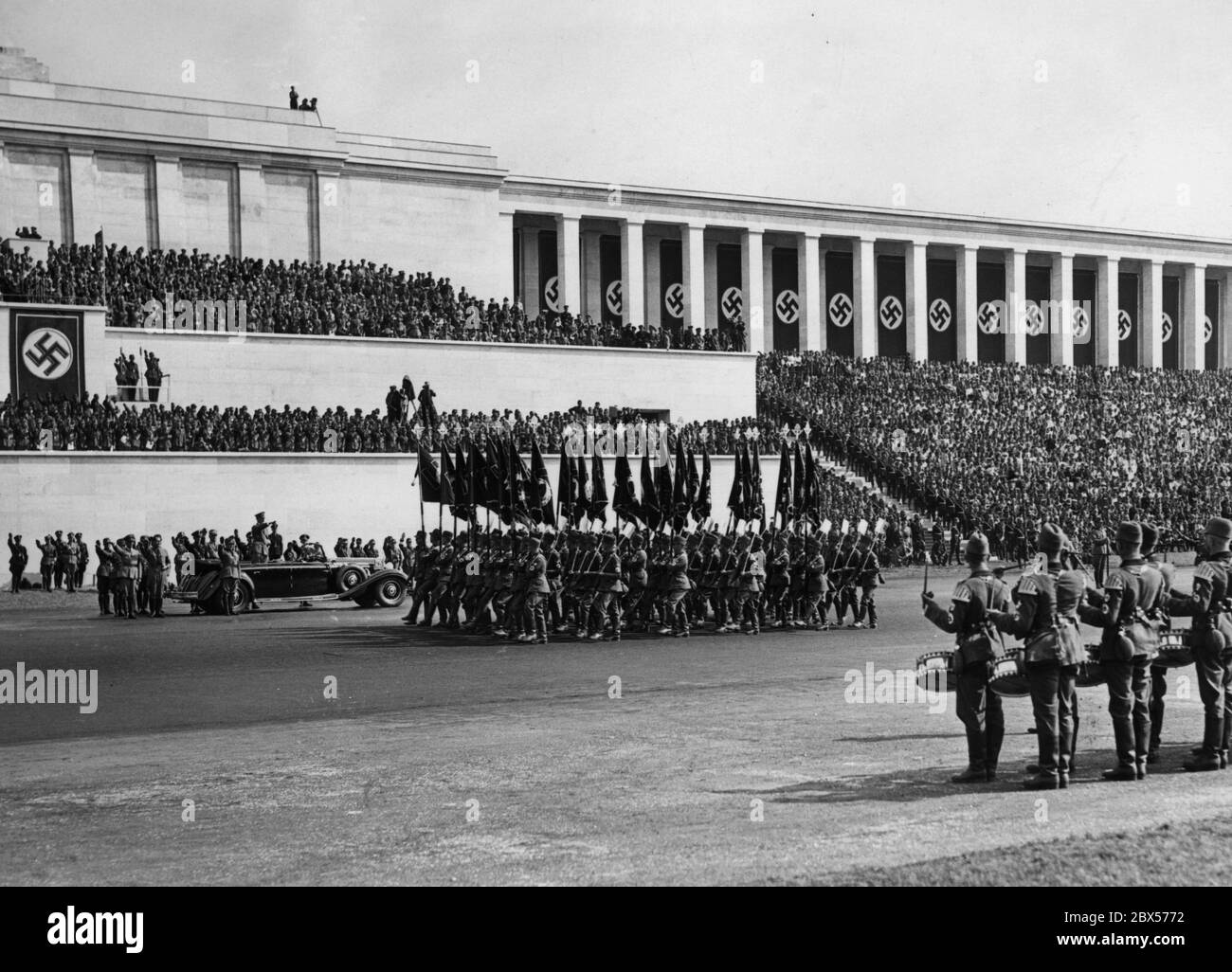 View of the march of formations of the Reich Labor Service with flags and spades on the Zeppelin Field past Adolf Hitler in his Mercedes and the grandstand of the Zeppelinfeld. Left in front of Hitler are Konstantin Hierl and Rudolf Hess. On the grandstand is a camera team. In the foreground, a drummer group of the RAD. Stock Photo