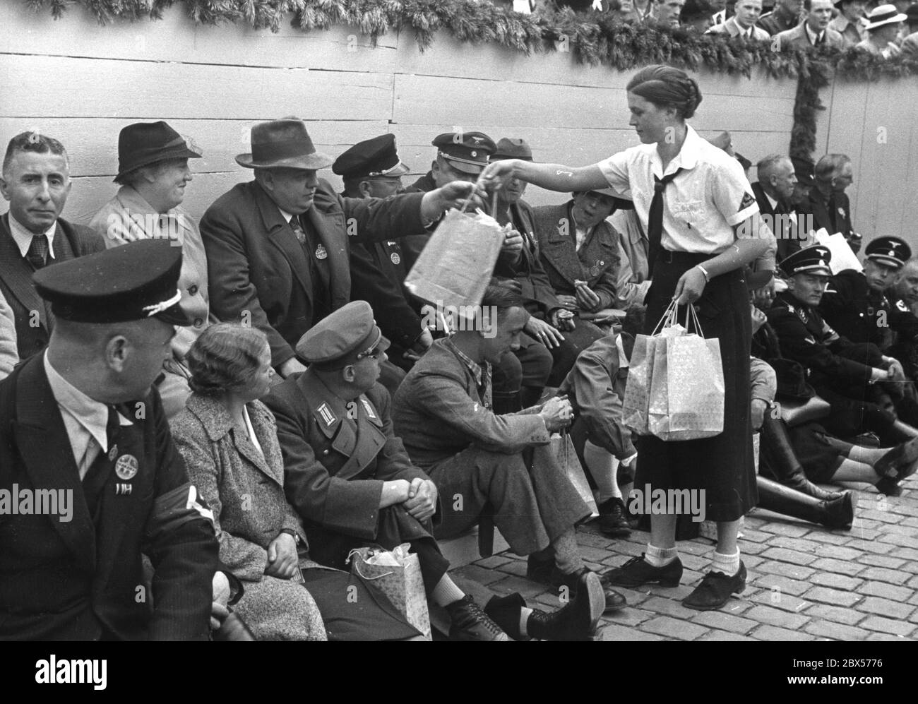 Young girls of the BDM give refreshments and bags with a small snack to former front soldiers in the VIP tribune during the parade of the Nazi organisations on the so-called Adolf-Hitler-Platz. Stock Photo
