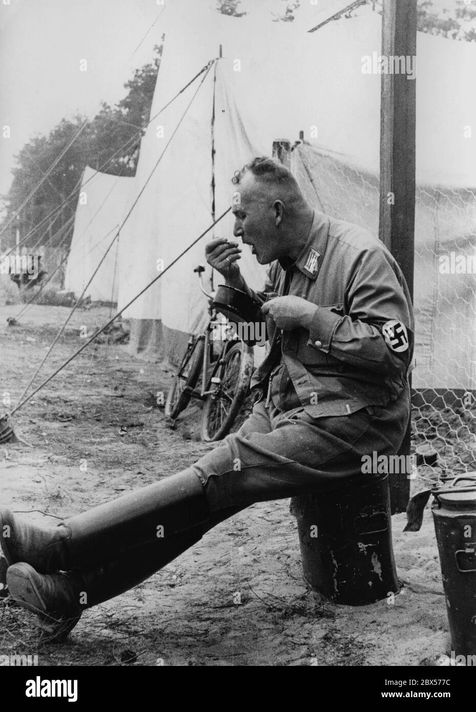 The Gau Saxony of the political leaders of the NSDAP set up their tents in the Langwasser camp during the Reich Party Congress. A party functionary is sitting on a thermos container and eats with his fingers from the lid of a cookware. A bicycle is leaning against the next tent. Stock Photo