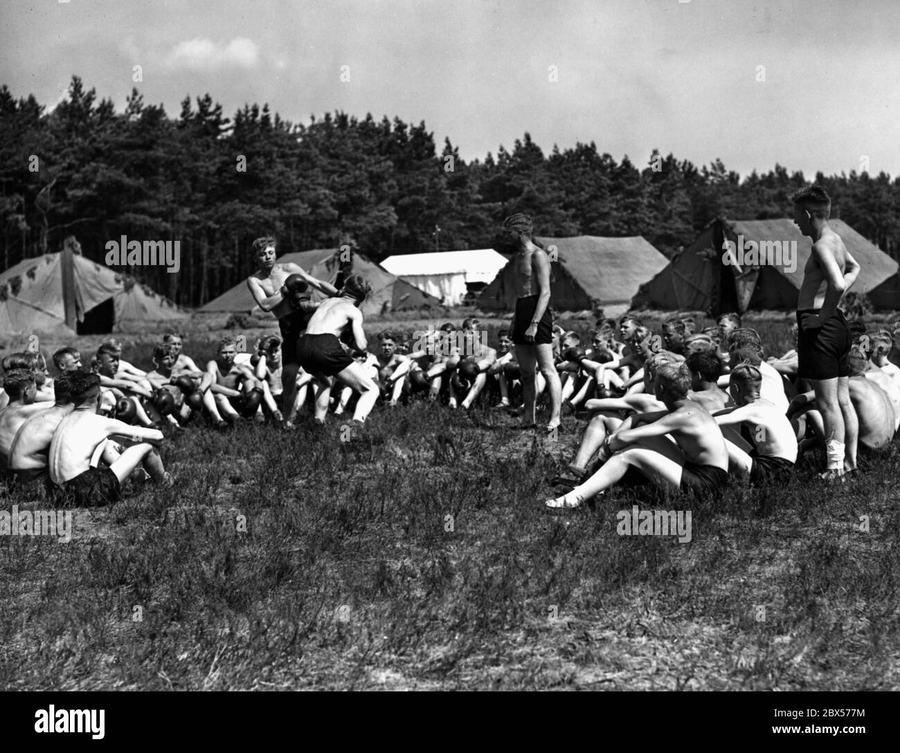 Motor Hitler Youth are boxing in the camp at "Potsdamer Platz" in the Nordoer Heide. Many other young people are sitting around them. In the background is the tent camp. Stock Photo