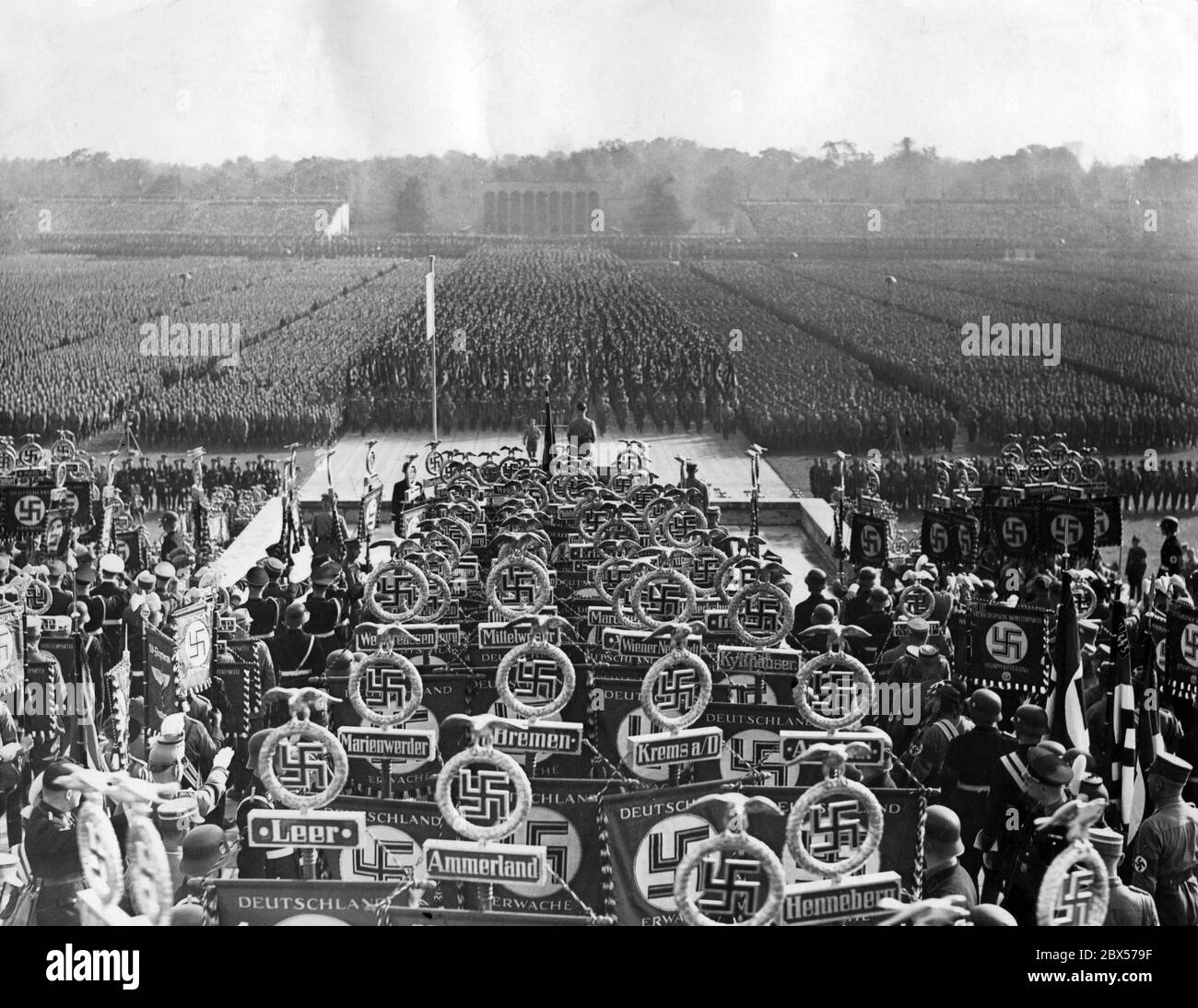 Overview of the grand roll call of SS, SA, NSKK and NSFK in the Luitpold Arena on the Nuremberg Nazi Party Rally Grounds during the flag march. Adolf Hitler is standing on the pulpit. In the foreground are standards of the SS and SA (Leer, Ammerland, Henneberg, Krems, Marienwerder, Bremen, Wiener Neustadt, Kyffhaeuaser, Mittelweser), in the background the Ehrenhalle (Hall of Honour). Stock Photo