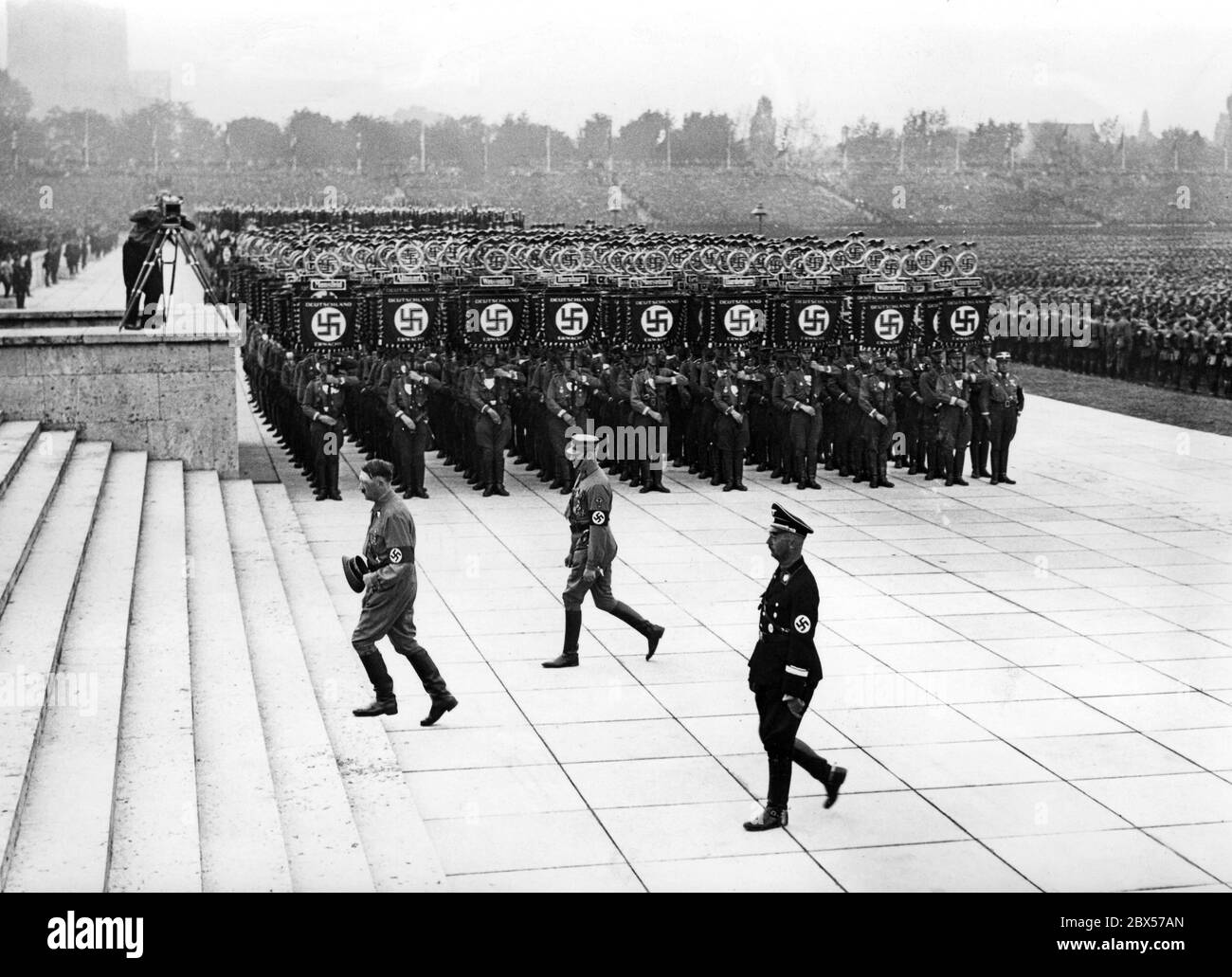 At the grand roll call of the SA, SS, NSFK and NSKK in Luitpoldhain, Adolf Hitler, Viktor Lutze and Heinrich Himmler walk to the platform from the front, past the standards of the SA and a cameraman. Stock Photo