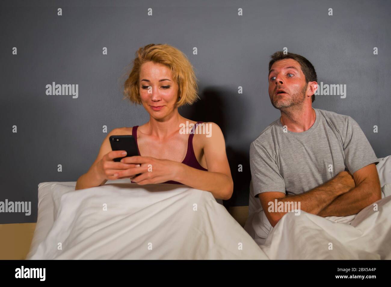 girlfriend or wife using mobile phone in bed and suspicious frustrated husband or boyfriend feeling upset suspecting betrayal and cheating in man woma Stock Photo