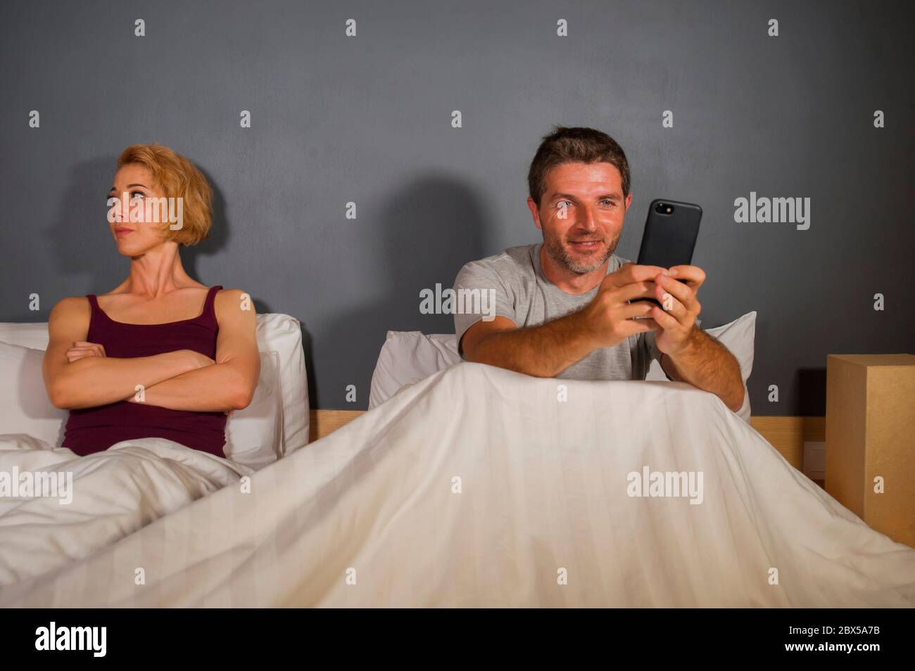 husband or boyfriend using mobile phone in bed and suspicious frustrated wife or girlfriend feeling upset suspecting betrayal and cheating in man woma Stock Photo
