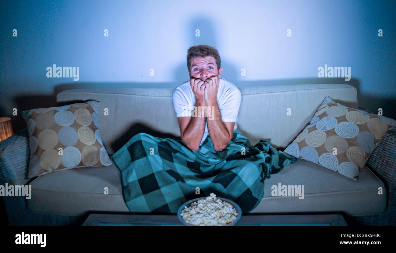 lifestyle portrait of attractive scared and nervous man watching suspense horror movie on television feeling stressed covering with blanket in panic s Stock Photo