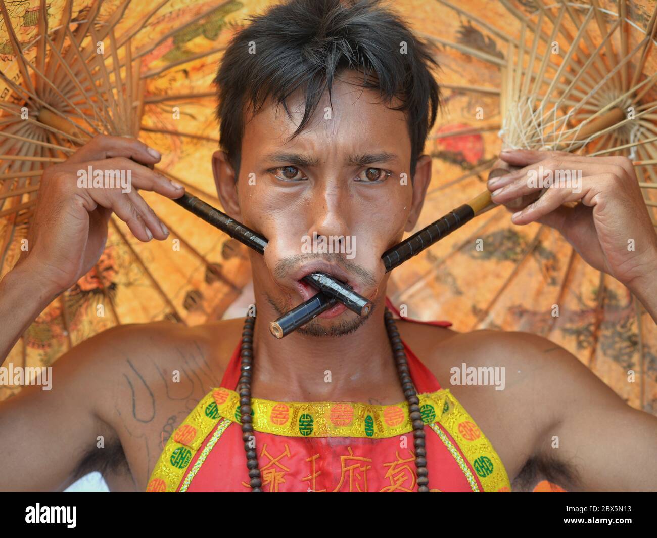 Thai Chinese Taoist devotee pierces his cheeks with two straight parasol handles during the Phuket Vegetarian Festival (Nine Emperor Gods Festival). Stock Photo