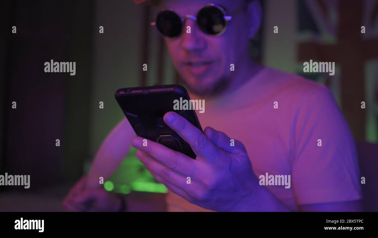 Hookah smoker using mobile phone. Handsome Caucasian Man in sunglasses sits in neon backlit. Selective focus on male hand holding phone in foreground Stock Photo