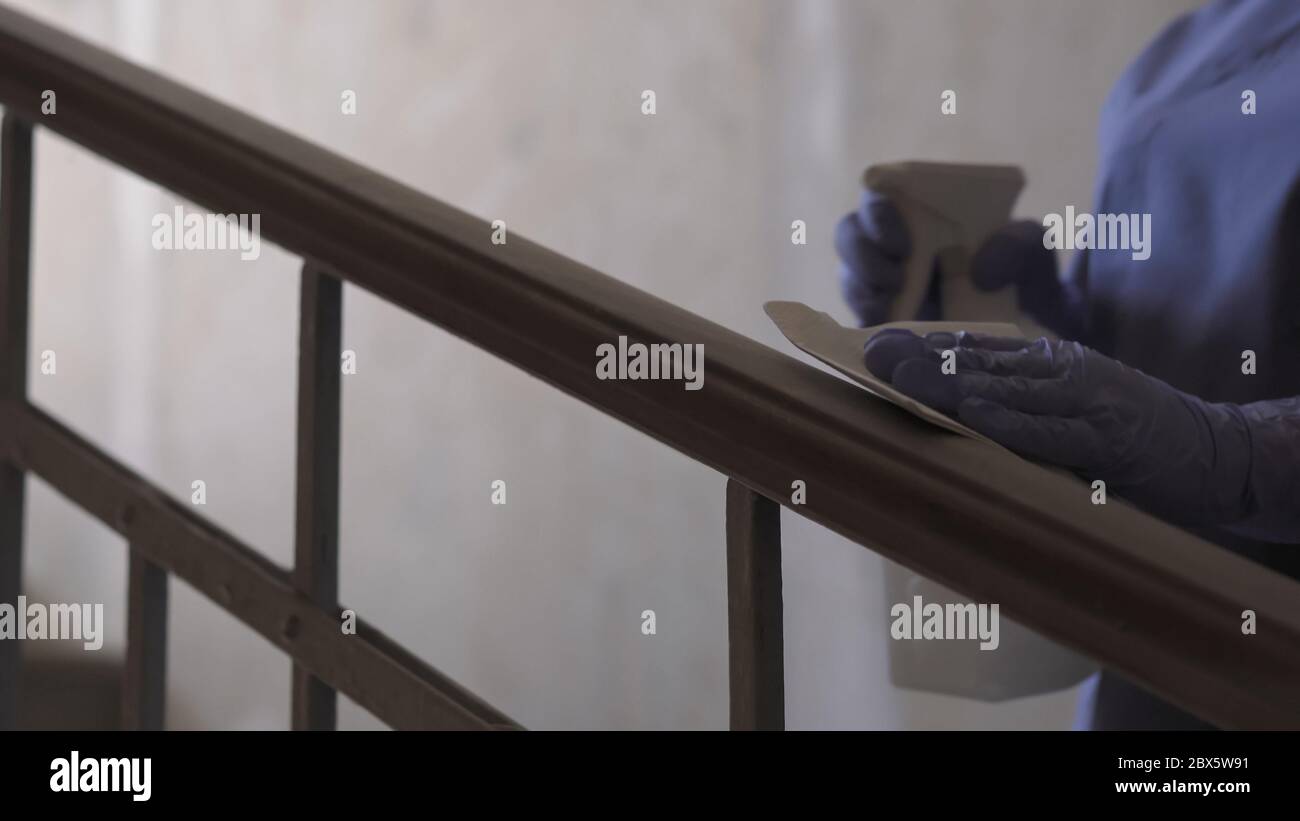 Worker wipes a liquid disinfectant railing stairwells. Employee in blue uniform treats public spaces with antibacterial or antiseptic liquid. Close up Stock Photo