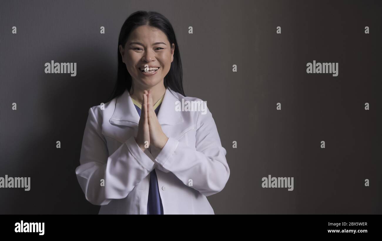 Happy doctor laughs joyfully clasped her hands together while standing against the gray wall. Beautiful brunette in medical uniform rejoices at the Stock Photo
