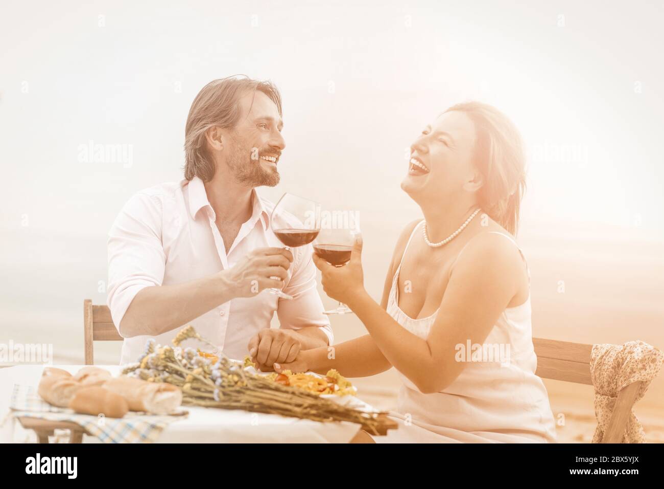 Happy man and woman drink wine in beach cafe. Lovely couple laughing holding hands and enjoying life while sitting together outdoors. Romantic midlife Stock Photo