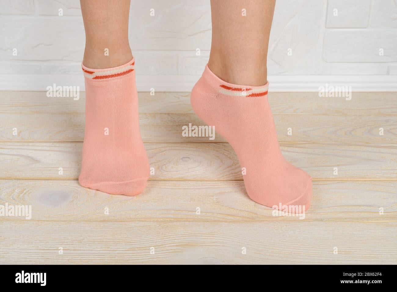 Woman feet in short pink cotton socks standing on toes on a white wood floor. Stand on tiptoe in low rise sports socks close-up. Front view. Stock Photo