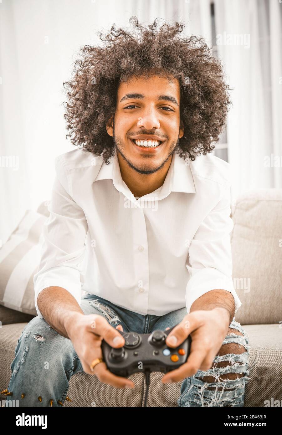 Happy Arab man smiles broadly playing computer game. Shaggy gamer holds Play Station joystick sitting at comfortable sofa at home. Funny time concept Stock Photo