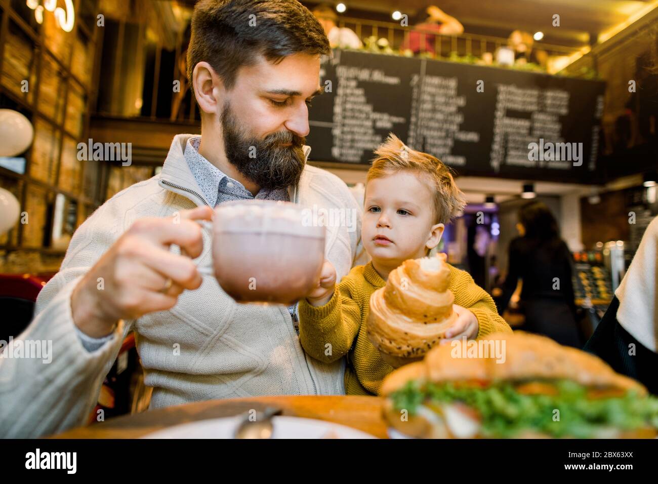 Happy handsome bearded Caucasian man, father, sitting at the table with his little son and holsing cup of latte or cocoa drink. Little cute boy Stock Photo