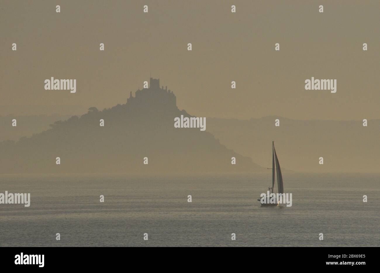 Misty atmospheric view of St Michaels Mount with an early start for a sailing yacht,Mounts Bay, Penzance, Cornwall, UK Stock Photo