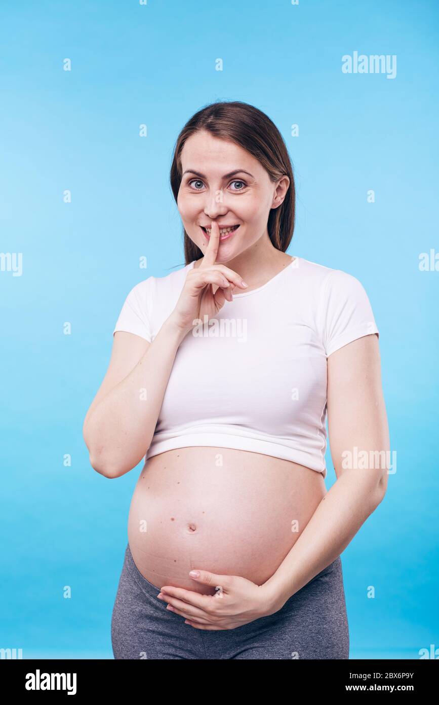 Happy young active woman keeping hand on bottom of her pregnant belly with  forefinger by mouth making shh gesture in isolation Stock Photo - Alamy