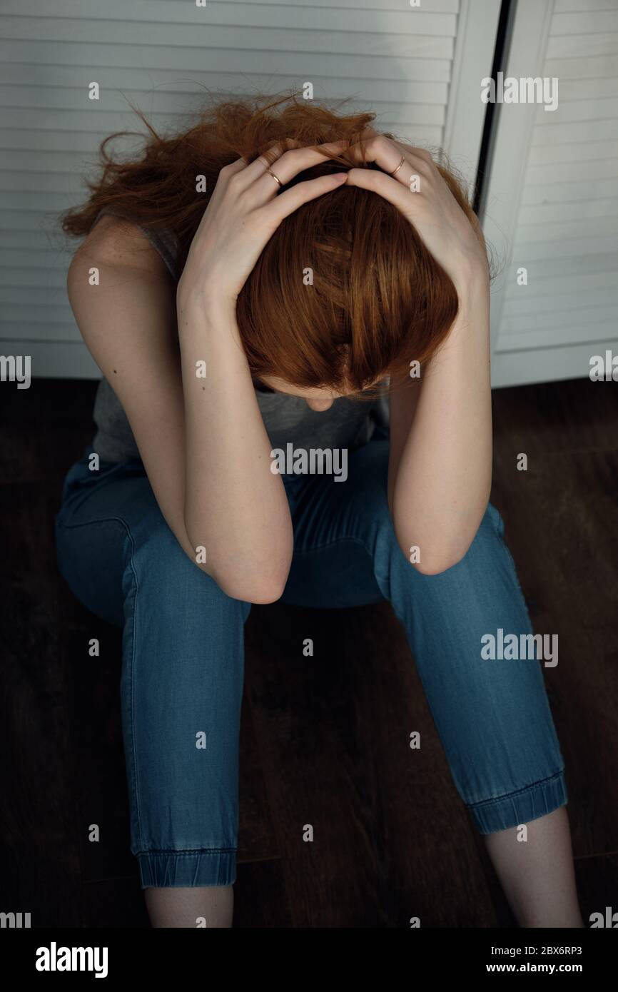 A red-haired girl in a t-shirt and pants sits on the floor with her head down and fingers in her hair, frame from above. Stock Photo