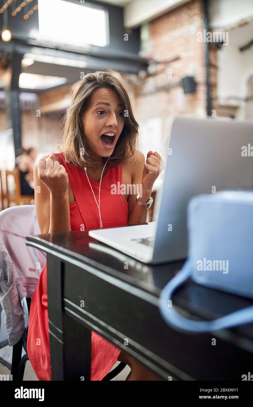 delighted young female watching content on her laptop with headphones in ears and fists up in YES position Stock Photo