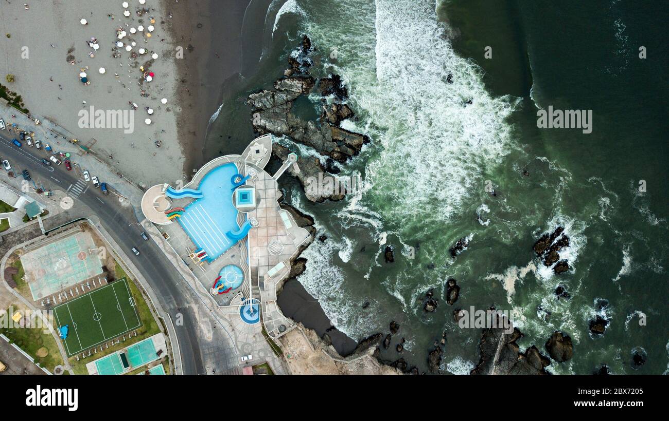 Aerial photo of an outdoor swimming pool at the coastline next to the rocks and crashing waves. Stock Photo