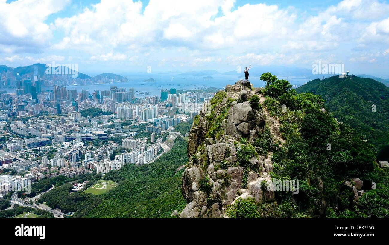 Aerial view on a solo traveling girl standing on my top of the rocky mountain with Hong Kong city and harbor in the background. Stock Photo