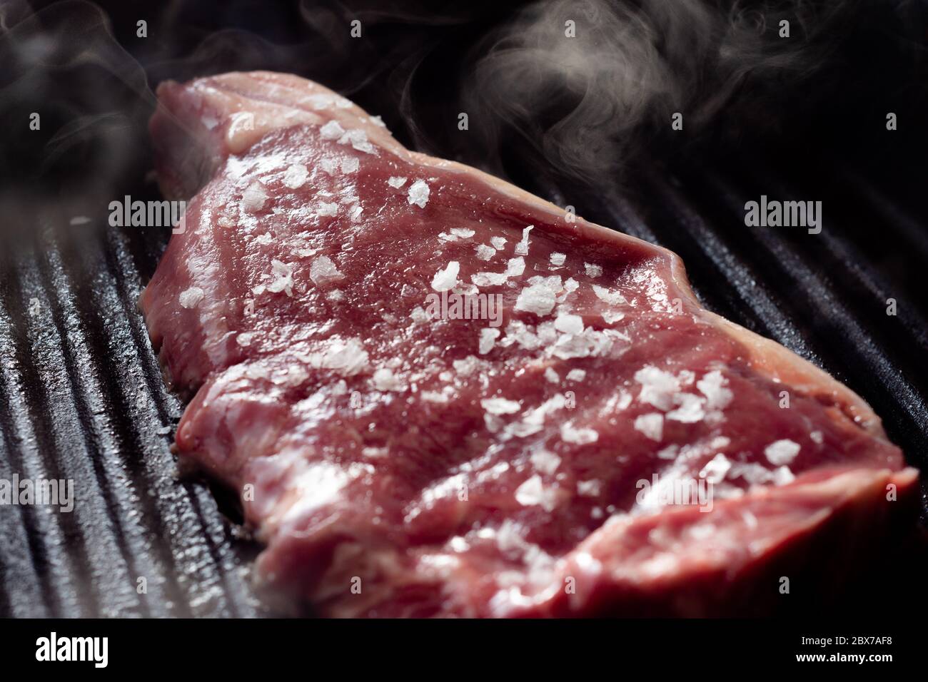 A slice of sirloin steak frying in a griddle pan. The steak is seasoned
