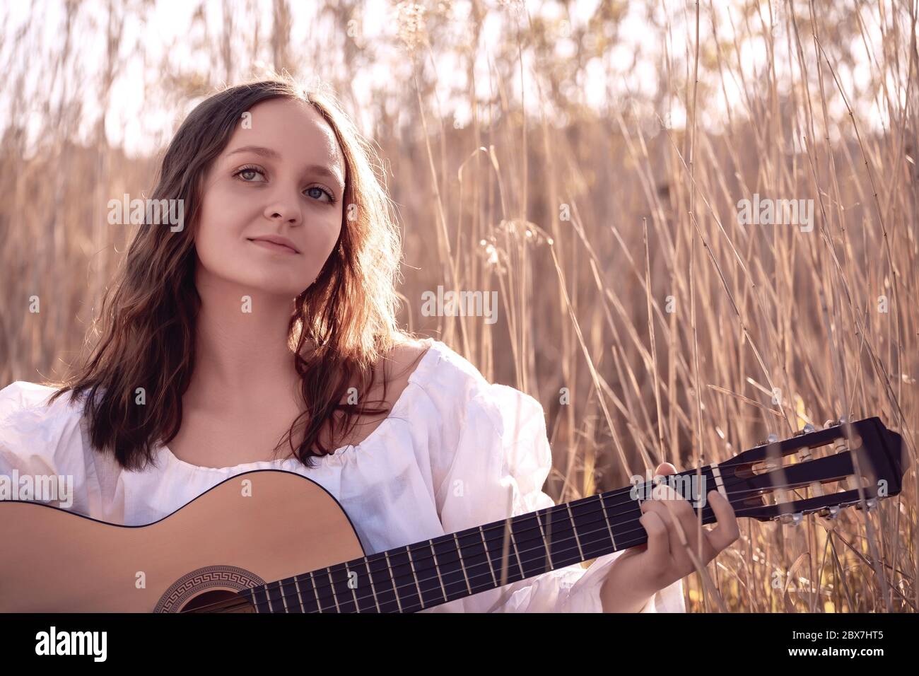 Young beautiful woman with long hair musician playing acoustic guitar at sunset field. Stock Photo