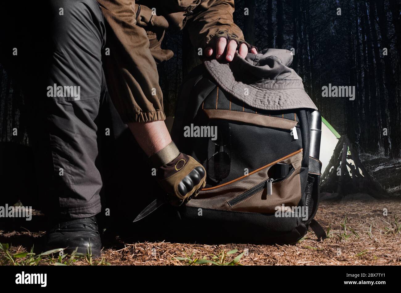 Man in tactical outfit holding a knife and kneeling for backpack with camping and tactical gear on night forest background. Stock Photo