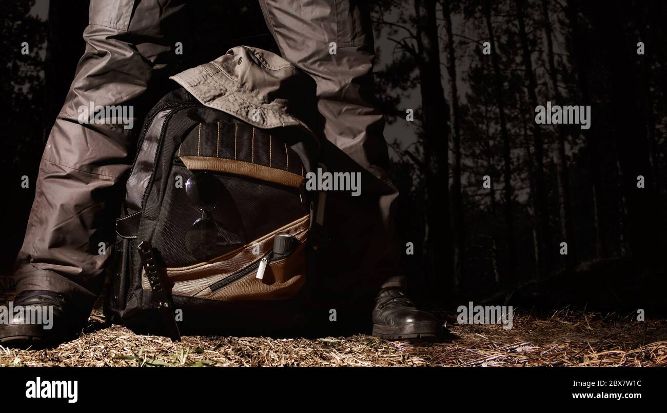 Man in tactical outfit standing over a backpack with camping and tactical gear on night forest background. Stock Photo
