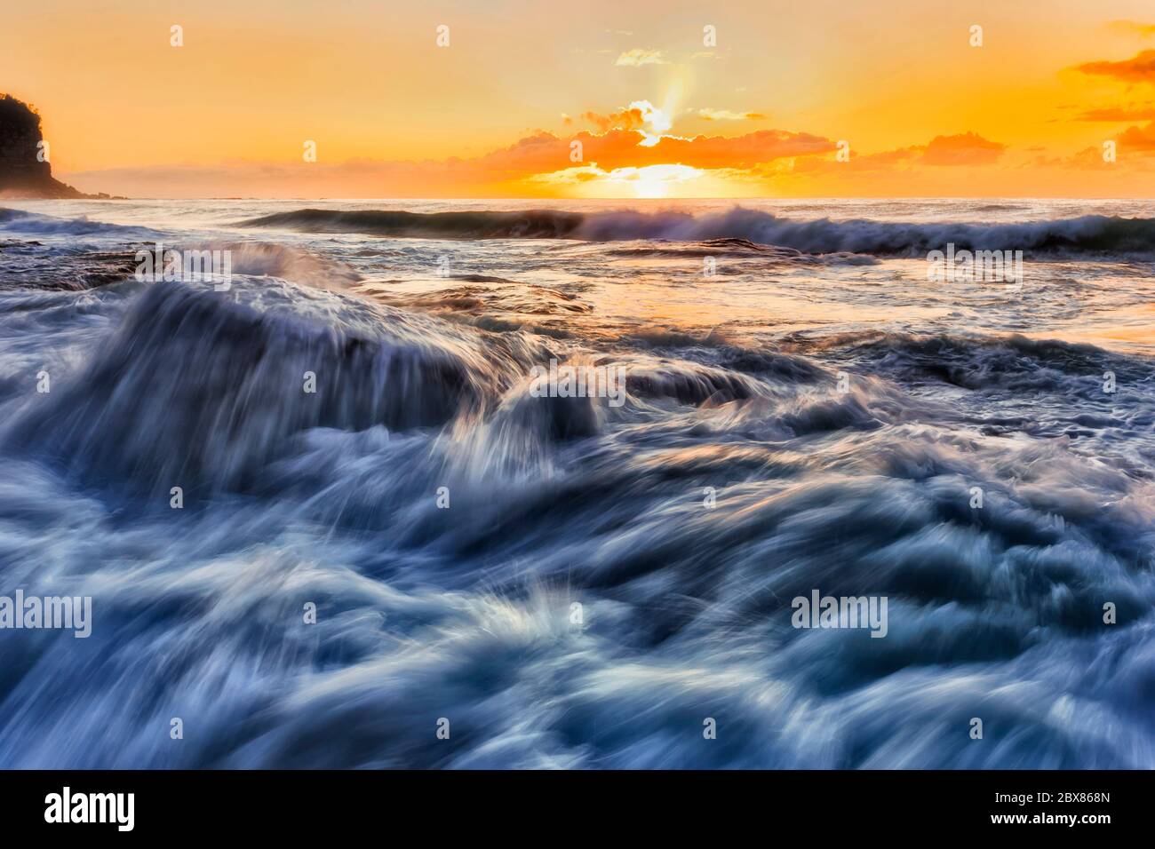 Stormy waves of Pacific ocean at sunrise against hot orange sun rollling over rocks in NOrthern beaches of Sydney. Stock Photo