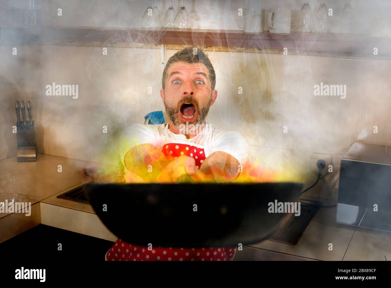 disaster home cook at kitchen- young funny and desperate man in cooking apron holding pan in flames in stress and fear making a mess of fire and smoke Stock Photo