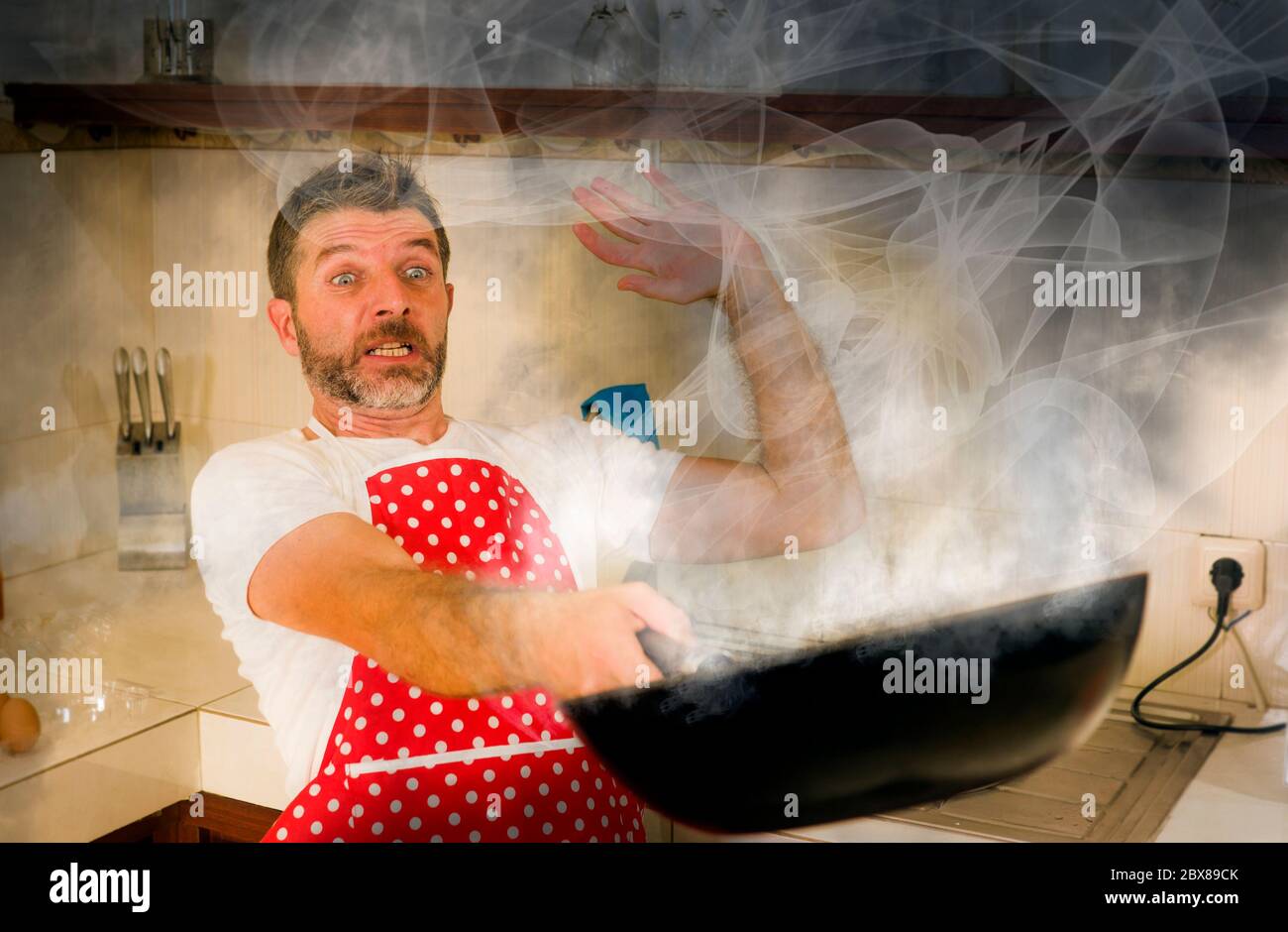 disaster home cook at kitchen- young funny and desperate man in cooking  apron holding pot in flames in stress and fear making a mess of fire and  smoke with food burning Stock