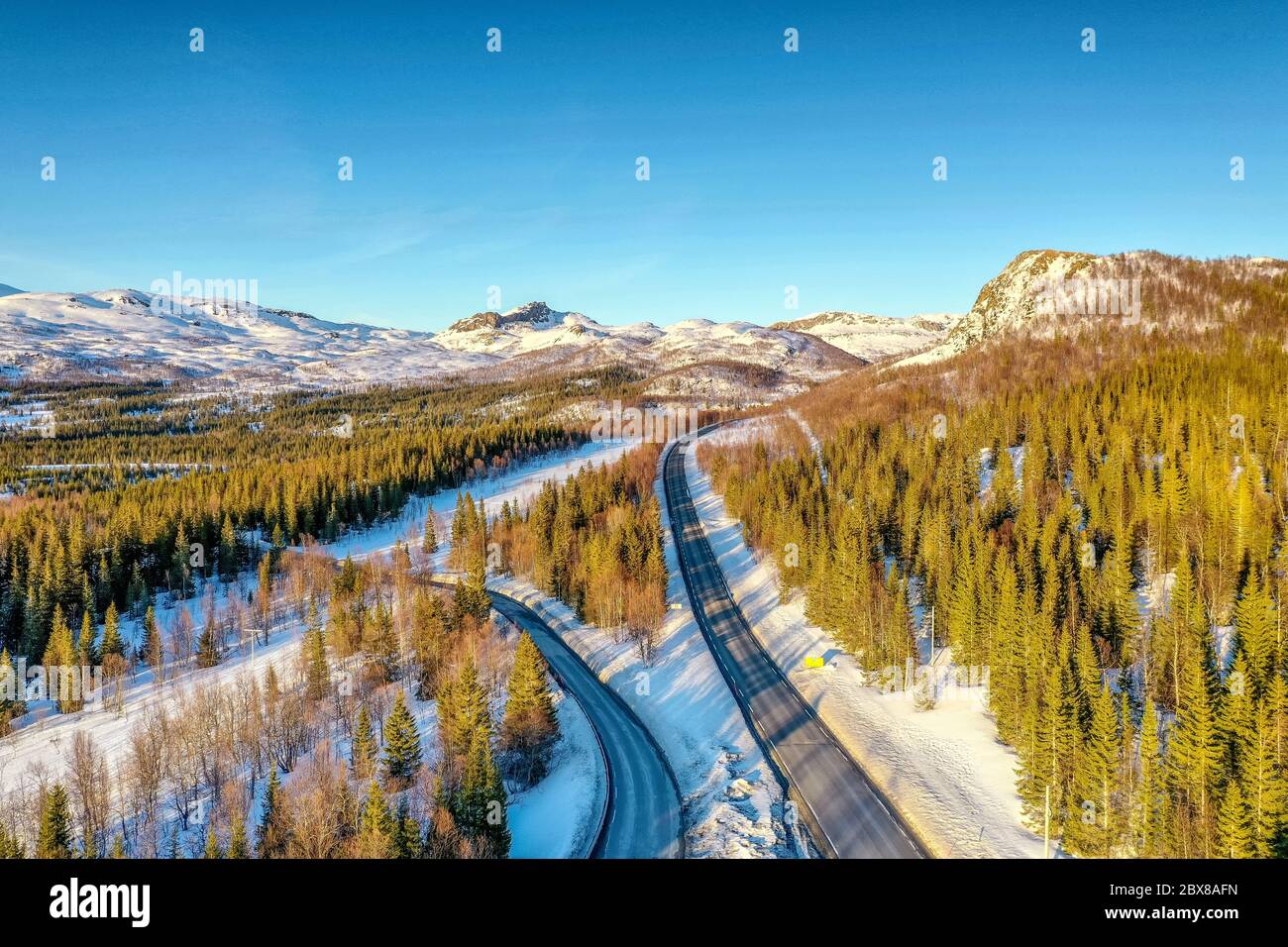 AERIAL: Sunset light over Norwegian snowy winter mountains with birche and pine trees, two roads, early spring, calm blue skies. Rossvatnet lake area, Stock Photo