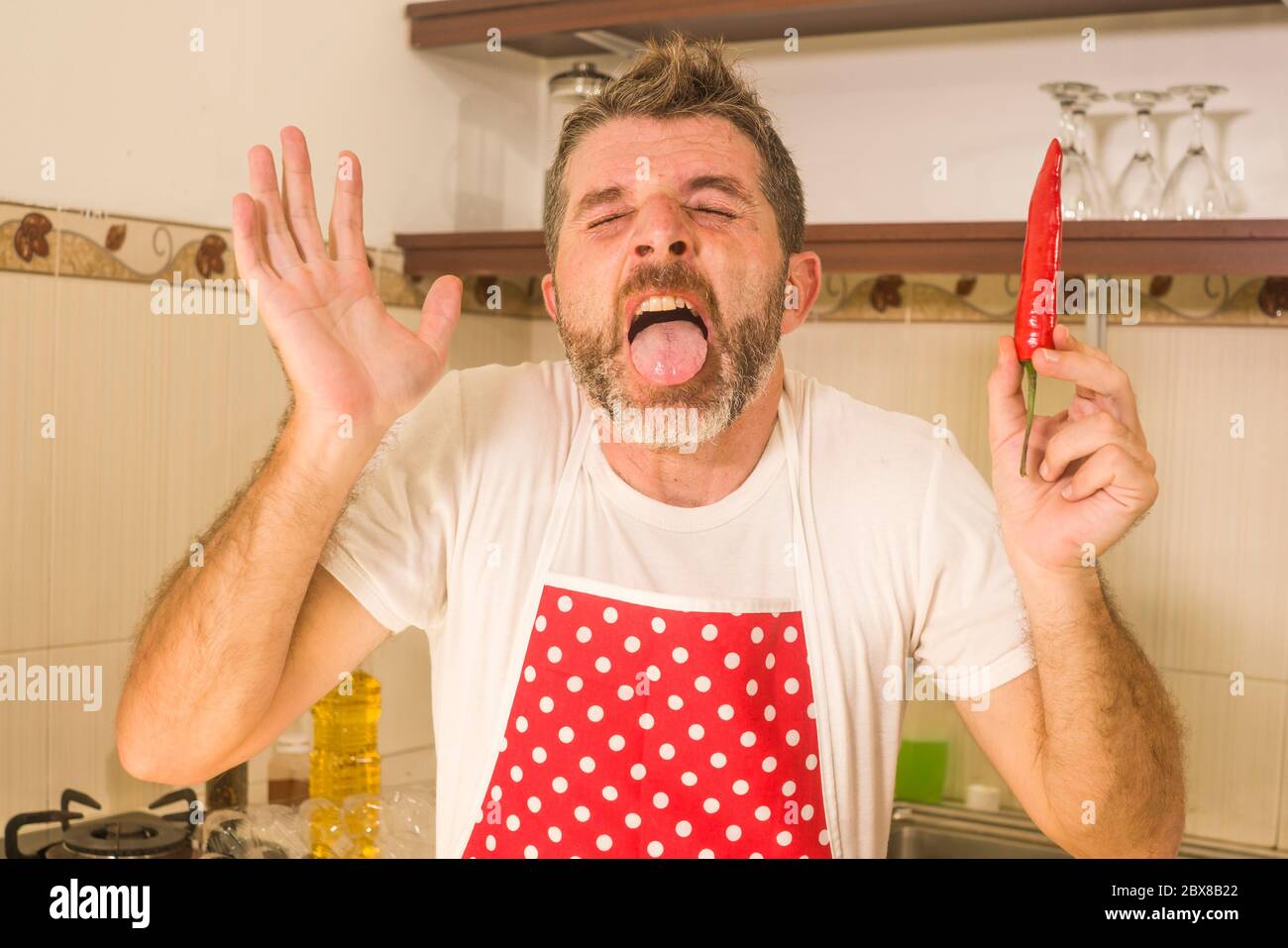 funny home portrait of home cook man in red kitchen apron holding spicy chili gesturing desperate taking his burning tongue out after tasting it crazy Stock Photo
