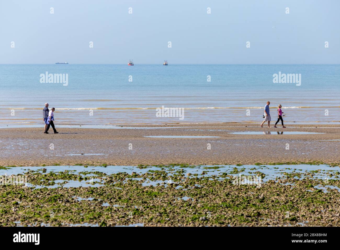 Worthing, Sussex, UK; 25th May 2020; Two Couples Walking Along The Seashore.  Two Fishing Boats and a Larger Ship Can Be Seen on the Horizen Stock Photo