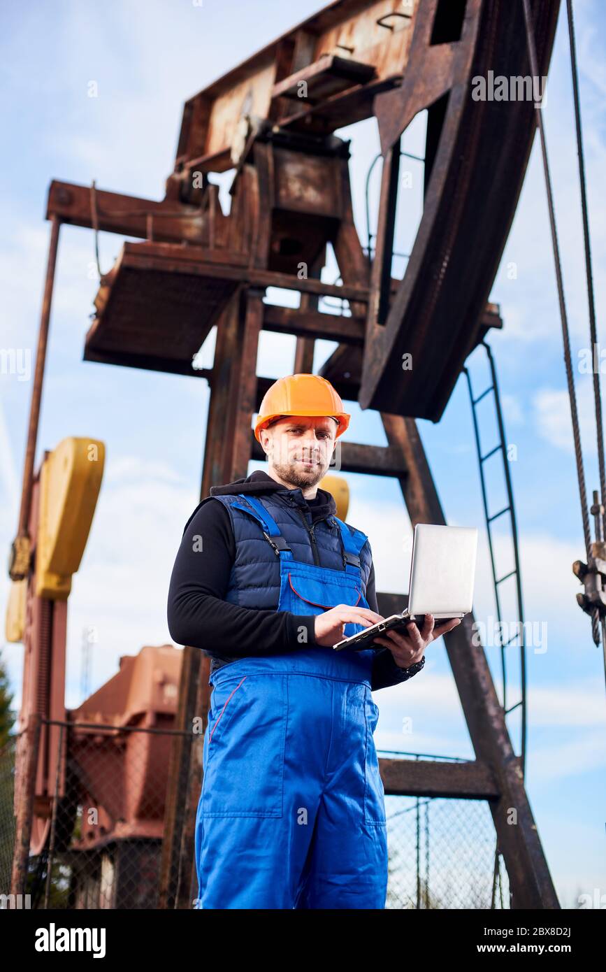 Vertical portrait of oil man, wearing blue overalls and orange helmet, with a laptop, standing with his back to an oil rig, checking oil pumping unit, making notes in his computer Stock Photo