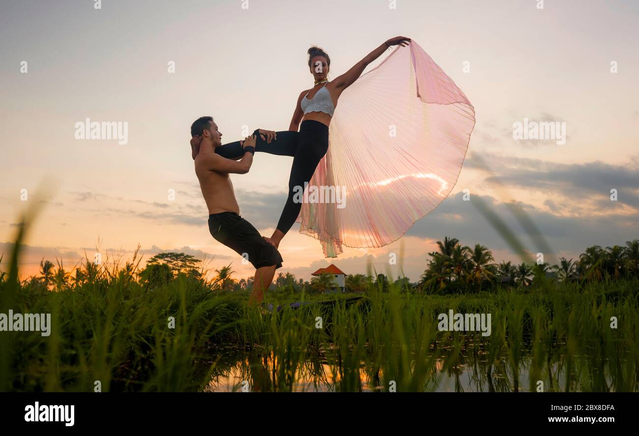 outdoors sunset acroyoga workout - young happy and fit couple practicing acro yoga drill at beautiful rice field enjoying nature and healthy lifestyle Stock Photo