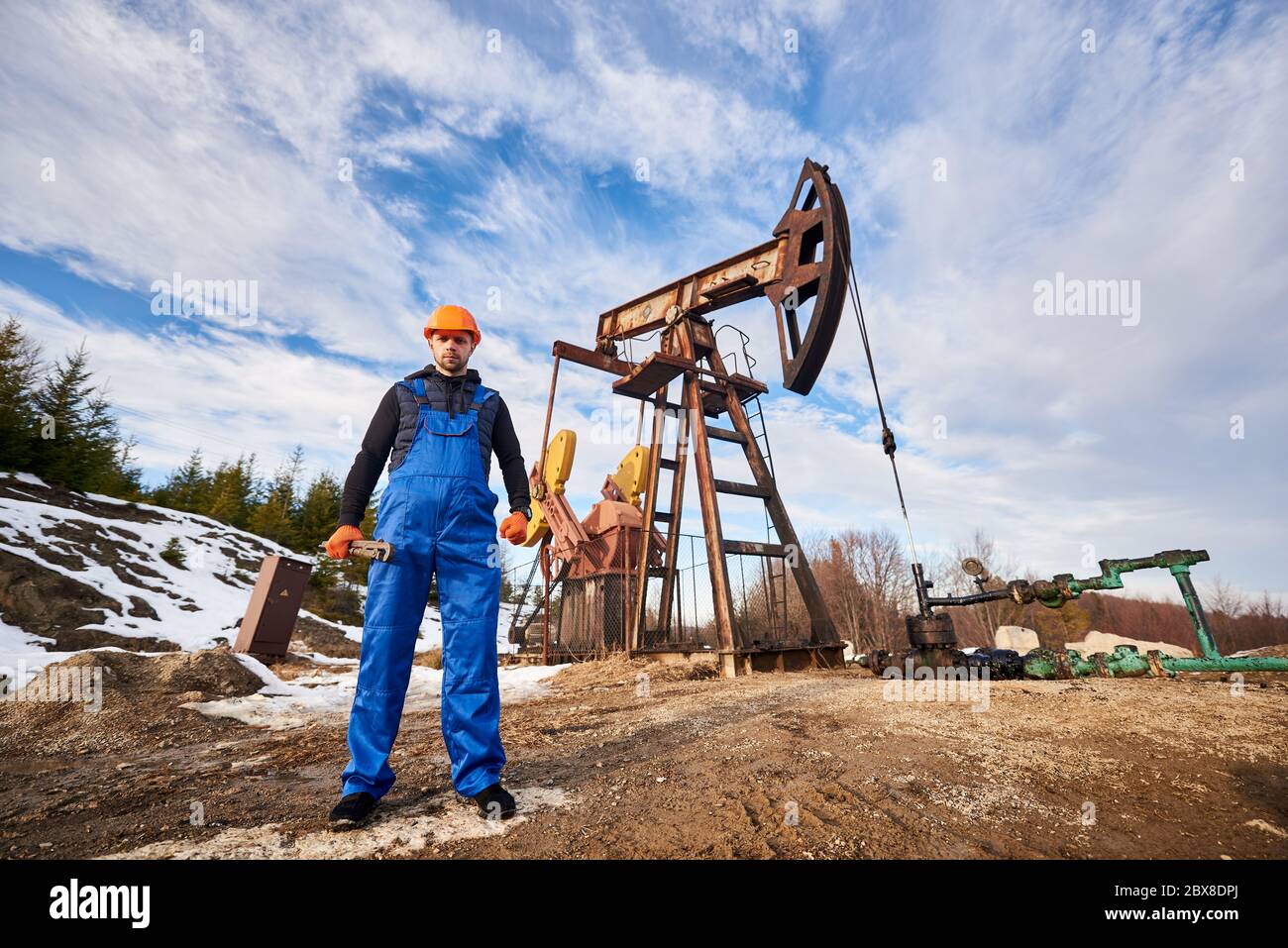 Oil worker during his working day in oil field, man wearing blue overalls and orange helmet, holding pipe wrench, looking at camera under beautiful sky. Concept of oil extraction, petroleum industry. Stock Photo