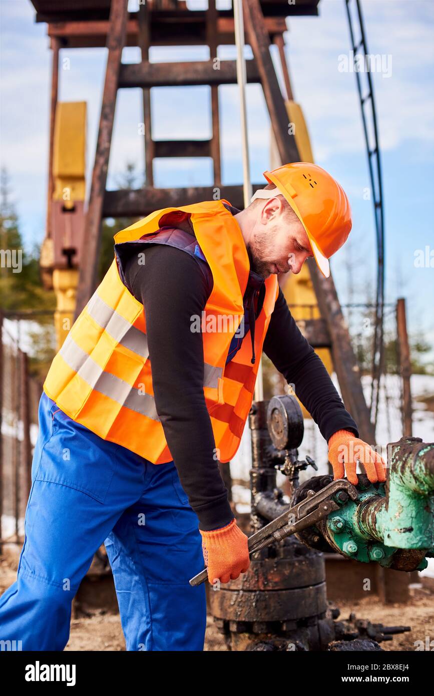 Side view of male technician using industrial wrench while repairing pipe of oil well pump jack. Oil worker in protective helmet and work vest tightening bolt on pipe. Concept of petroleum industry Stock Photo