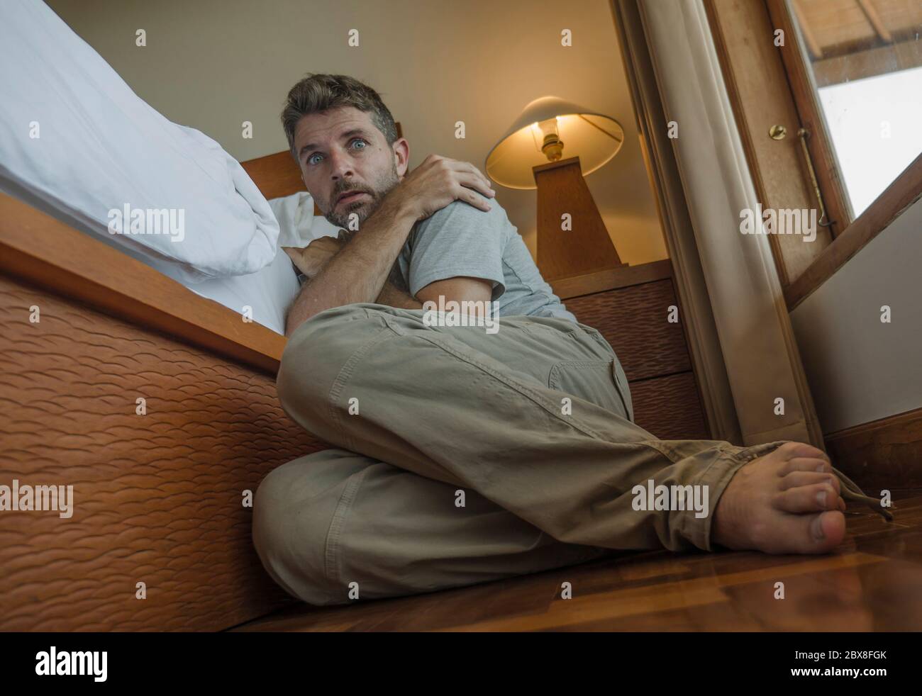 dramatic home portrait of young depressed and desperate man sitting on bedroom floor next to bed suffering depression and anxiety feeling overwhelmed Stock Photo