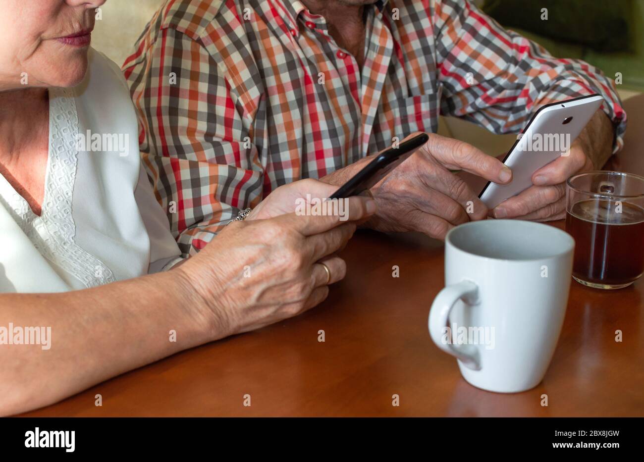 Elderly couple using modern mobile phones at home while enjoying cups of hot drinks. Concept of elderly people and technology, personal development... Stock Photo
