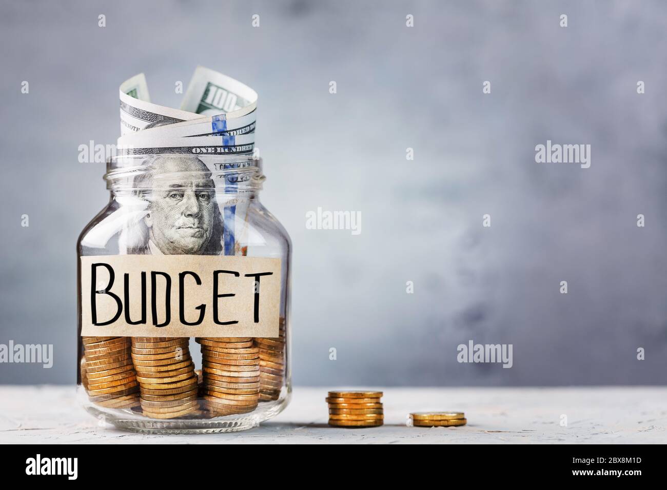 Glass Jar with gold coins, with a hundred-dollar bill and a sticker saying Budget on a gray background with free space for text. Stock Photo